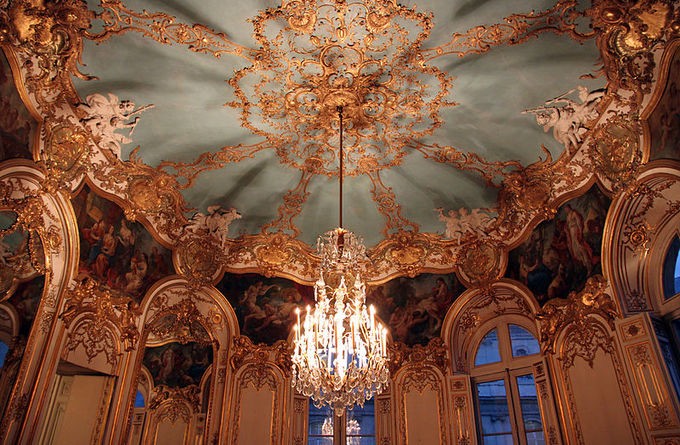 Chandelier hanging from a blue and gold ceiling in a room lined with arched windows and mosaics